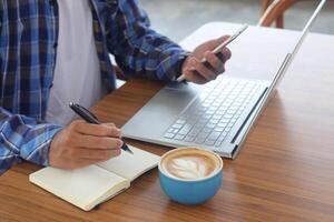 vicino su di maschio mano scrittura su Nota libro con penna, mostrando vuoto schermo di il computer portatile e smartphone. Lavorando nel bar concetto con un' tazza di caffè foto