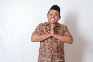 ritratto di sorridente asiatico uomo indossare batik camicia e songkok mostrando scusarsi e benvenuto mano gesto. Ramadan pubblicità concetto. isolato Immagine su grigio sfondo foto