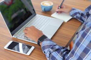 vicino su di maschio mano scrittura su Nota libro con penna, mostrando vuoto schermo di il computer portatile e smartphone. Lavorando nel bar concetto con un' tazza di caffè foto