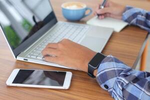 vicino su di maschio mano scrittura su Nota libro con penna, mostrando vuoto schermo di il computer portatile e smartphone. Lavorando nel bar concetto con un' tazza di caffè foto