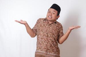 ritratto di confuso asiatico uomo indossare batik camicia e songkok con attraversato mani, puntamento di lato, fabbricazione scelta, la scelta fra Due oggetti. isolato Immagine su grigio sfondo foto