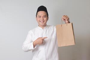 ritratto di asiatico musulmano uomo nel bianca koko camicia con zucchetto dando mestiere carta shopping Borsa con regalo dentro. isolato Immagine su grigio sfondo foto