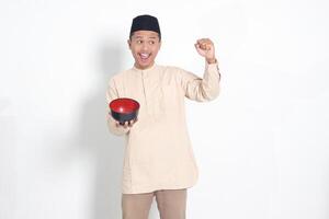 ritratto di eccitato asiatico musulmano uomo nel koko camicia con zucchetto mostrando e Tenere un vuoto piatto. ciotola modello per cibo marca. isolato su bianca sfondo foto