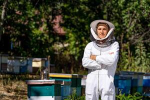 uomo apicoltore sta mani incrociate vicino orticaria. maschio nel protettivo bianca uniforme circondato con verde alberi nel il giardino. foto