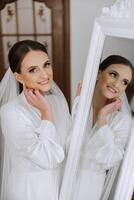 nel il mattina, un incredibilmente bellissimo e lussuoso sposa, vestito nel un' bianca veste, pose di il specchio nel sua Camera da letto. mattina di il sposa con un' lussuoso acconciatura e naturale trucco. foto