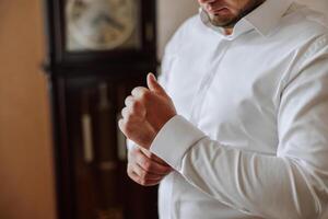 un' uomo nel un' bianca camicia sta di il finestra nel il camera e si allaccia il pulsanti su il suo collare e maniche. orologio su mano. elegante attività commerciale ritratto di un' uomo, avvicinamento foto. il sposo è preparazione. foto