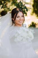 un' brunetta sposa nel un' voluminoso bianca vestito detiene un' mazzo, pose Tenere sua riccioli. il velo è gettato in il aria. bellissimo capelli e trucco. nozze foto sessione nel natura.