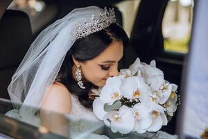 il sposa è seduta nel un' nero auto con un' mazzo di fiori nel sua mani e sembra su di il macchina. ritratto di un' piuttosto timido sposa nel un' macchina. sposa, Sorridi, emozioni. foto