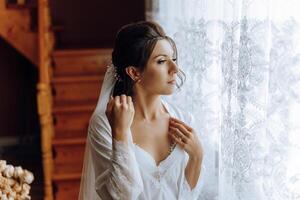 ritratto di un incredibilmente bellissimo ragazza sposa nel un' bianca accappatoio nel il Camera da letto, il sposa pose nel il mattina prima il nozze cerimonia. foto