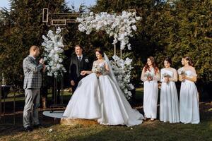 un' maestro di cerimonie dà un' discorso mentre un' sposa e sposo hold mani vicino un' bianca floreale arco foto