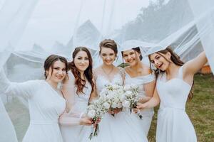 nozze fotografia nel natura. un' brunetta sposa e sua amiche posa sotto un' velo, Tenere mazzi di fiori foto