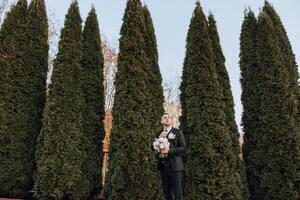 ritratto di un' bello giovane sposo su un autunno giorno all'aperto nel un elegante completo da uomo e Tenere un' nozze mazzo di fiori. all'aperto foto. bello uomo nel un' attività commerciale completo da uomo. foto