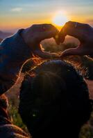 un' giovane ragazza fabbricazione cuore simbolo con sua mani a tramonto foto