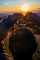 un' giovane ragazza fabbricazione cuore simbolo con sua mani a tramonto foto