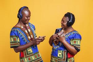 sorridente nero uomo e donna indossare etnico Abiti ascoltando per musica nel cuffia. allegro coppia nel relazione Tenere mobile telefoni e godendo elenco di riproduzione nel senza fili auricolari foto