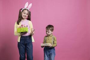 poco bambini Tenere fatto a mano cestino con dipinto uova su telecamera, giovane ragazza fare pace cartello e in posa con sua piccolo fratello. dolce fratelli sensazione gioioso di Pasqua celebrazione. foto