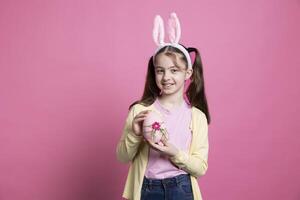 bello carino piccolo ragazza con coniglietto orecchie Spettacoli un' grande rosa uovo, in posa con Pasqua decorazioni e disposizioni. giovane allegro bambino essere Uscito di primavera celebrazione, fatto a mano arte. foto