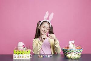 carino contento bambino piccolo pittura Pasqua uova con acquerello nel studio, decorazione fatto a mano festivo ornamenti contro rosa sfondo. giovane bambino gode creatività e mestiere, colorazione con pennello. foto