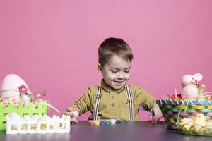 entusiasta giovane bambino utilizzando acquerelli per dipingere uova e decorazioni, la creazione di artistico progetti nel preparazione per Pasqua Domenica vacanza. allegro giovanotto godendo cravatta tintura colorazione nel studio. foto