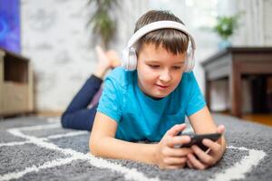 un' bambino ragazzo con Telefono e cuffie è dire bugie su il pavimento foto