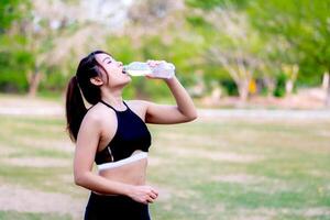 donna idratante dopo l'allenamento nel un' parco in mezzo verde, incarnando fitness e Salute. adolescenziale donna potabile acqua a partire dal un' acqua bottiglia. bellissimo ragazza nel nero esercizio Abiti. foto