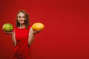 foto di un' carino ragazza nel un' rosso vestito sorridente con un' anguria e melone nel sua mani isolato su un' luminosa rosso sfondo