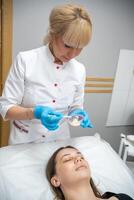 medico cosmetologo o dermatologo fabbricazione viso maschera nel cosmetologia salone. professionale estetista l'applicazione viso maschera su caucasico donna viso dire bugie su letto nel accappatoio. foto