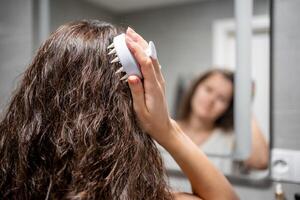 giovane donna con buio Riccio capelli fare se stesso capelli cuoio capelluto massaggio con cuoio capelluto massaggiatore o capelli spazzola per capelli crescita stimolante a casa foto