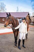 bella biondo professionale femmina fantino in piedi vicino Marrone cavallo su azienda agricola nel inverno. amicizia con cavallo concetto foto