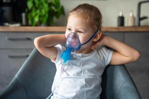 carino poco ragazza siamo seduta e Tenere un' nebulizzatore maschera pendente contro il viso, vie aeree trattamento concetto foto