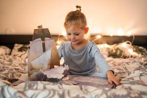 poco ragazza lettura pop su libro nel casa letto nel Natale ambiente con luci sfondo foto