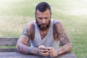 tatuato barbuto uomo scrive con inteligente Telefono nel il parco foto