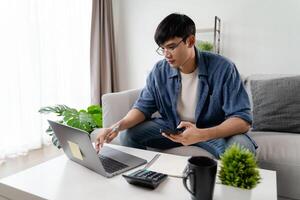 il uomo nel casuale Abiti Lavorando con un' computer portatile, computer, inteligente Telefono, calcolatrice seduta su il divano nel il vivente camera a casa, Lavorando a partire dal casa concetto. foto