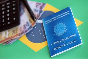 brasiliano opera carta e sociale sicurezza blu libro e reale i soldi fatture con calcolatrice e penna su bandiera di federativo repubblica di brasile foto