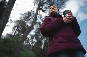 Visualizza a partire dal il parte inferiore di un' contento sorridente donna Tenere viaggio thermos tazza, riposo o escursioni a piedi nel il foresta foto