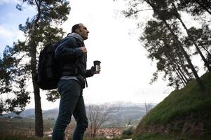 lato ritratto di un' giovane uomo con zaino e viaggio tazza, durante un' il trekking nel il foresta, montagne su il sfondo foto