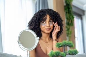 moderno cosmetologia e cerotti contro fatica sotto il occhi. allegro giovane africano americano donna l'applicazione loro sotto sua occhi. foto
