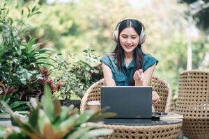 un' allegro giovane donna con cuffie si siede a un' malacca sedia, coinvolgente nel un' video chiamata su sua il computer portatile in mezzo lussureggiante verde. foto