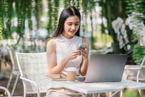 un' giovane donna è assorbito nel sms su sua smartphone, con un' il computer portatile e caffè a un all'aperto tavolo circondato di verde. foto