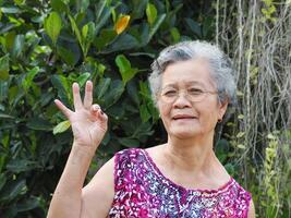 un' anziano asiatico donna con corto bianca capelli sorridente e guardare a il telecamera mentre in piedi nel un' giardino foto