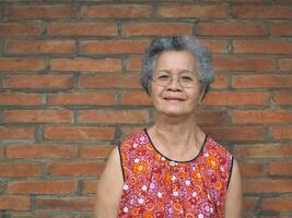 un anziano asiatico donna sorridente e guardare a il telecamera mentre in piedi con un' mattone parete sfondo. spazio per testo. concetto di anziano persone e assistenza sanitaria foto