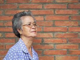 anziano asiatico donna sorridente e guardare lontano mentre in piedi con un' mattone parete sfondo. concetto di anziano persone e assistenza sanitaria. foto