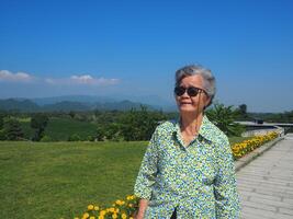 anziano donna indossare occhiali da sole, sorridente e guardare a il telecamera mentre in piedi nel il tè piantagione. spazio per testo. concetto di anziano persone e rilassamento foto