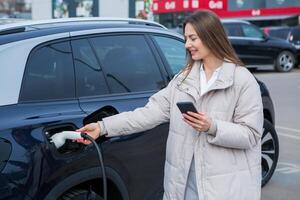 giovane donna ricarica sua elettrico auto a un' ricarica stazione nel il città. eco carburante concetto. il concetto di l'ambiente amichevole trasporto. ricarica batteria a partire dal ricarica stazione. foto