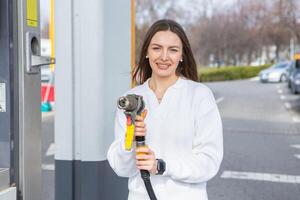 giovane donna Tenere un' carburante ugello nel sua mano mentre rifornimento carburante auto a gas stazione. un' fermare per rifornimento carburante a il gas stazione. fare il pieno il auto con gas. foto