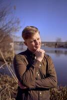 ritratto di un' giovane uomo nel un' Marrone camicia su il banca di un' primavera fiume foto