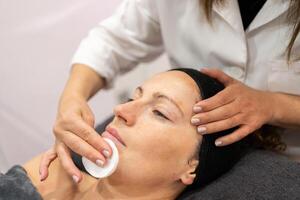 Ritaglia estetista pulizia viso di cliente con pelle cura cotone tampone nel bellezza salone foto