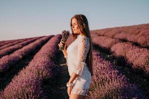 donna lavanda campo. contento spensierato donna nel un' bianca vestito a piedi nel un' lavanda campo e odorare un' lavanda mazzo su tramonto. ideale per caldo e ispirazione concetti nel voglia di girovagare e viaggio. foto