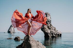 donna viaggio mare. giovane contento donna nel un' lungo rosso vestito in posa su un' spiaggia vicino il mare su sfondo di vulcanico rocce, piace nel Islanda, condivisione viaggio avventura viaggio foto