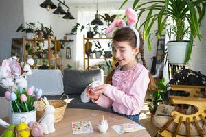 un' carino ragazza con rosa coniglietto orecchie fa un Pasqua mestiere - decora un uovo nel il modulo di un' unicorno con strass, corno, fiori nel il interno di un' Casa con impianti. foto